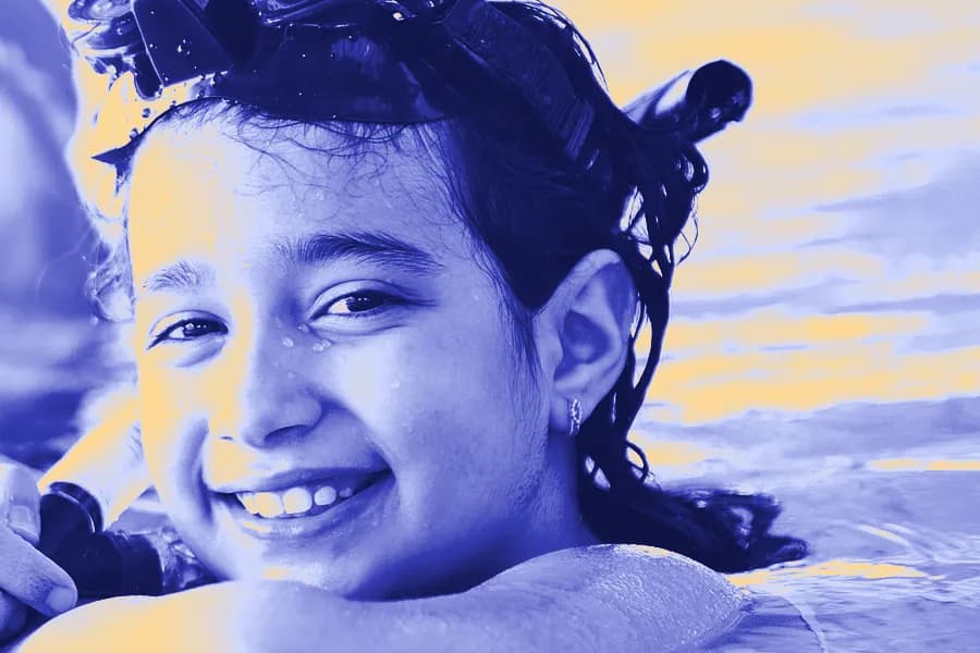 A smiling girl with swim goggles pushed up on her head rests at the edge of a pool.