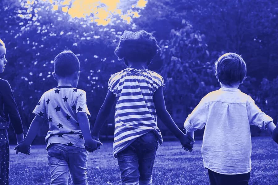 Young children walking hand in hand away from the camera across a grassy park.