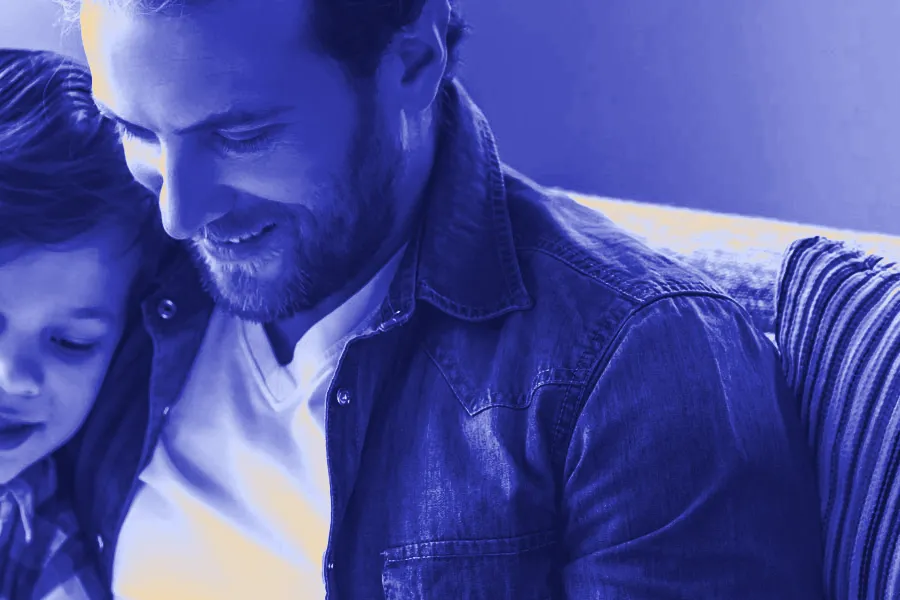 A smiling father in a denim shirt sits on a sofa and looks down at a book with his young son.
