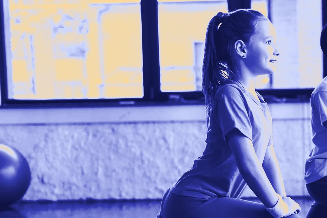A smiling girl in a T-shirt stretches on the floor of a sunny gym.