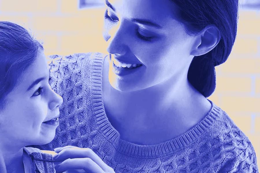 A smiling mother leans in close to her young daughter, who smiles back at her.