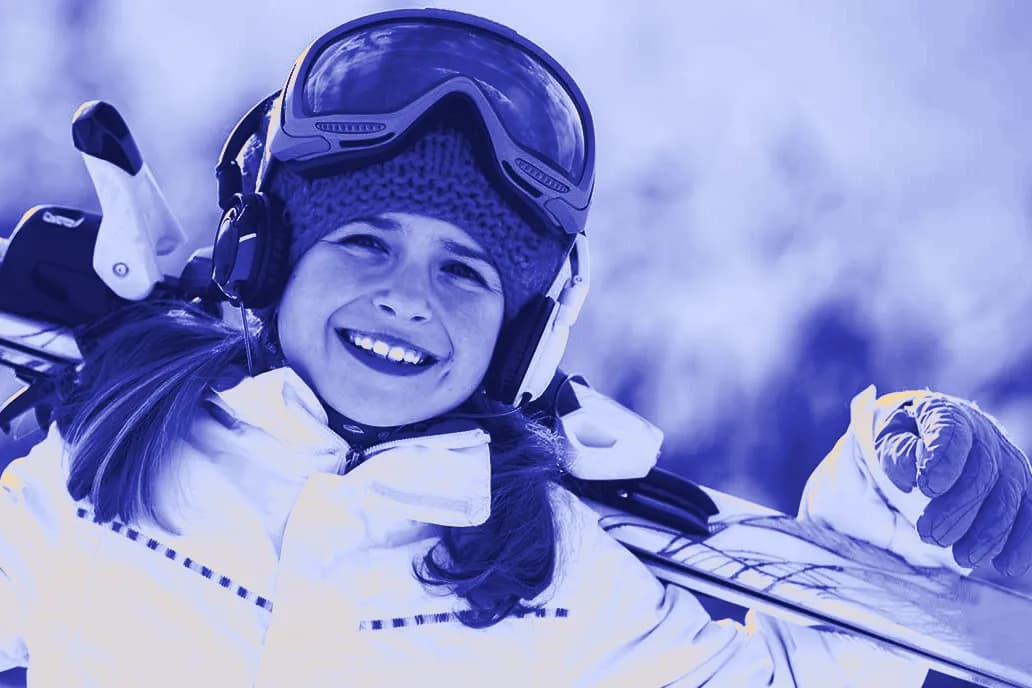 A smiling girl in a ski jacket, knit hat and goggles carrying her skis.