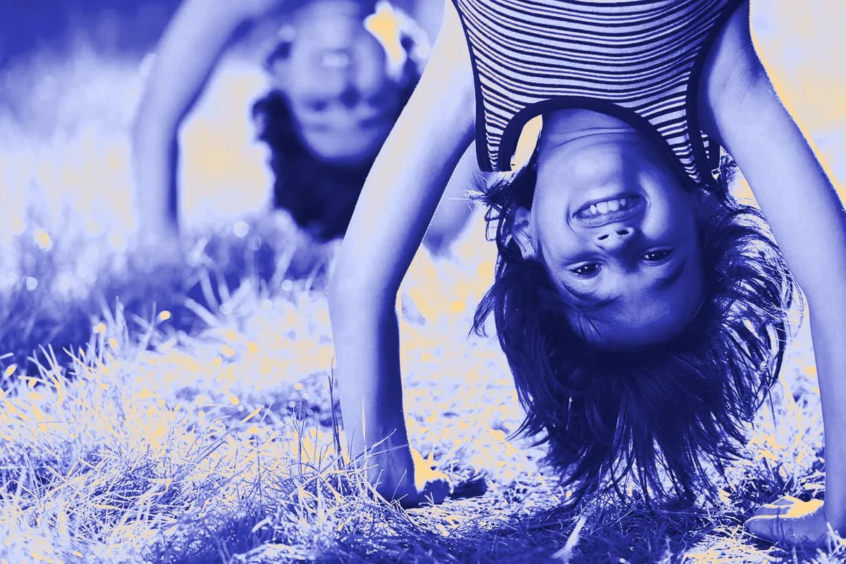 Two children playing in the grass, one bent over and smiling upside down at the camera.