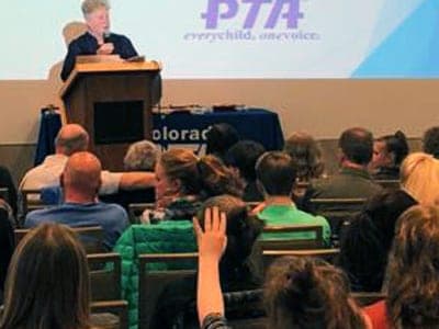 A speaker at a podium addresses a seated audience in front of a PTA slide.